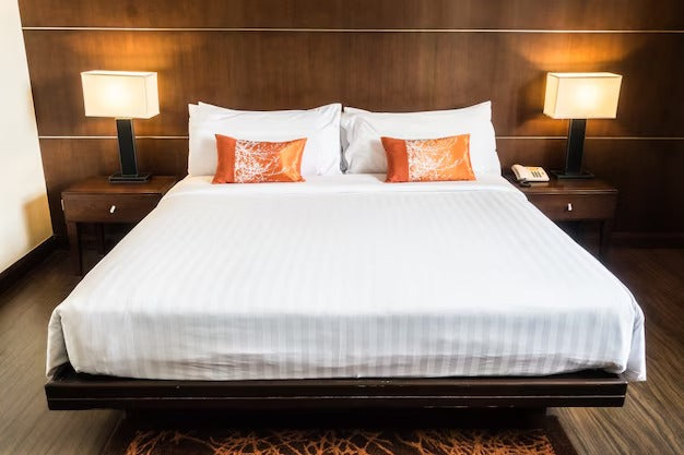 Modern hotel room with a neatly made double murphy bed, white bedding, orange cushions, wooden headboard, and matching side tables with lamps.