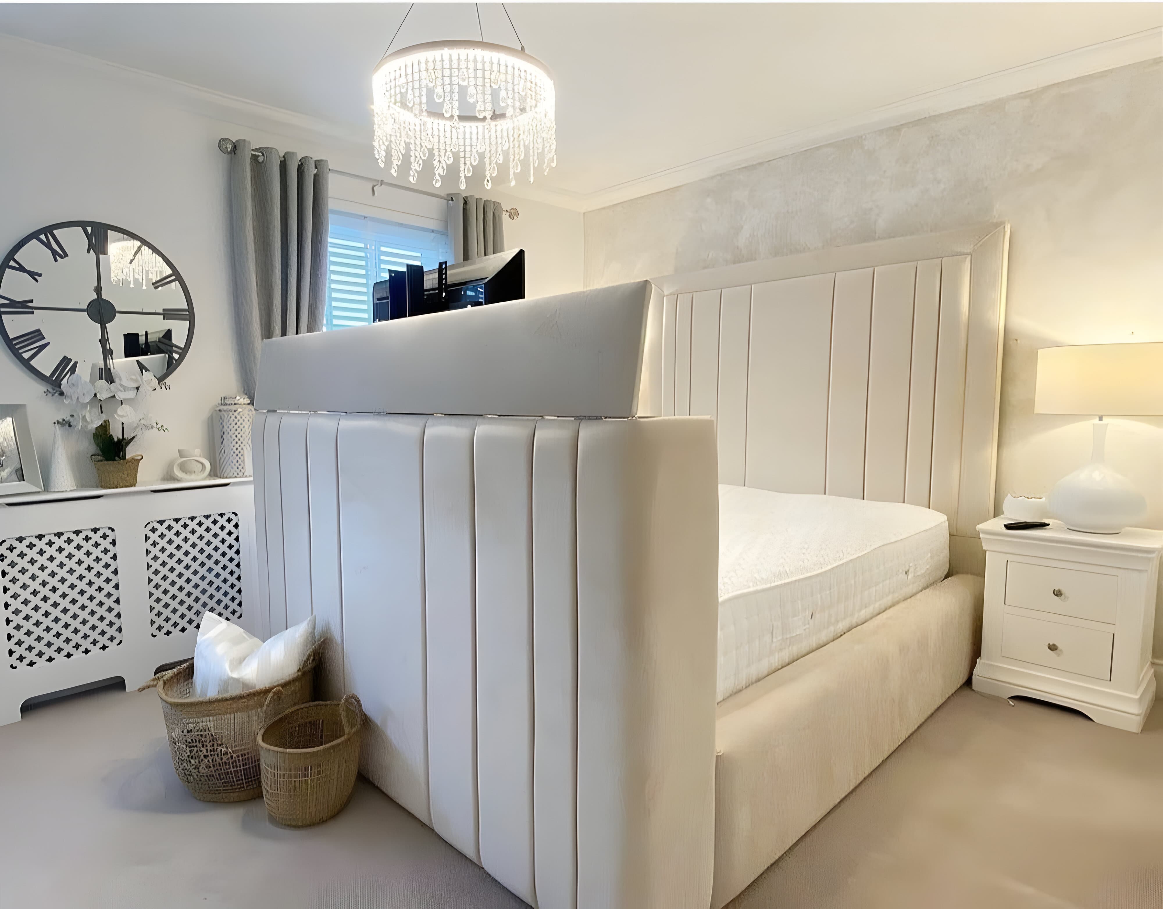 A bedroom with a double TV bed featuring a beige upholstered headboard and footboard, a TV emerging from the footboard, a large wall clock, and a white nightstand with a lamp.