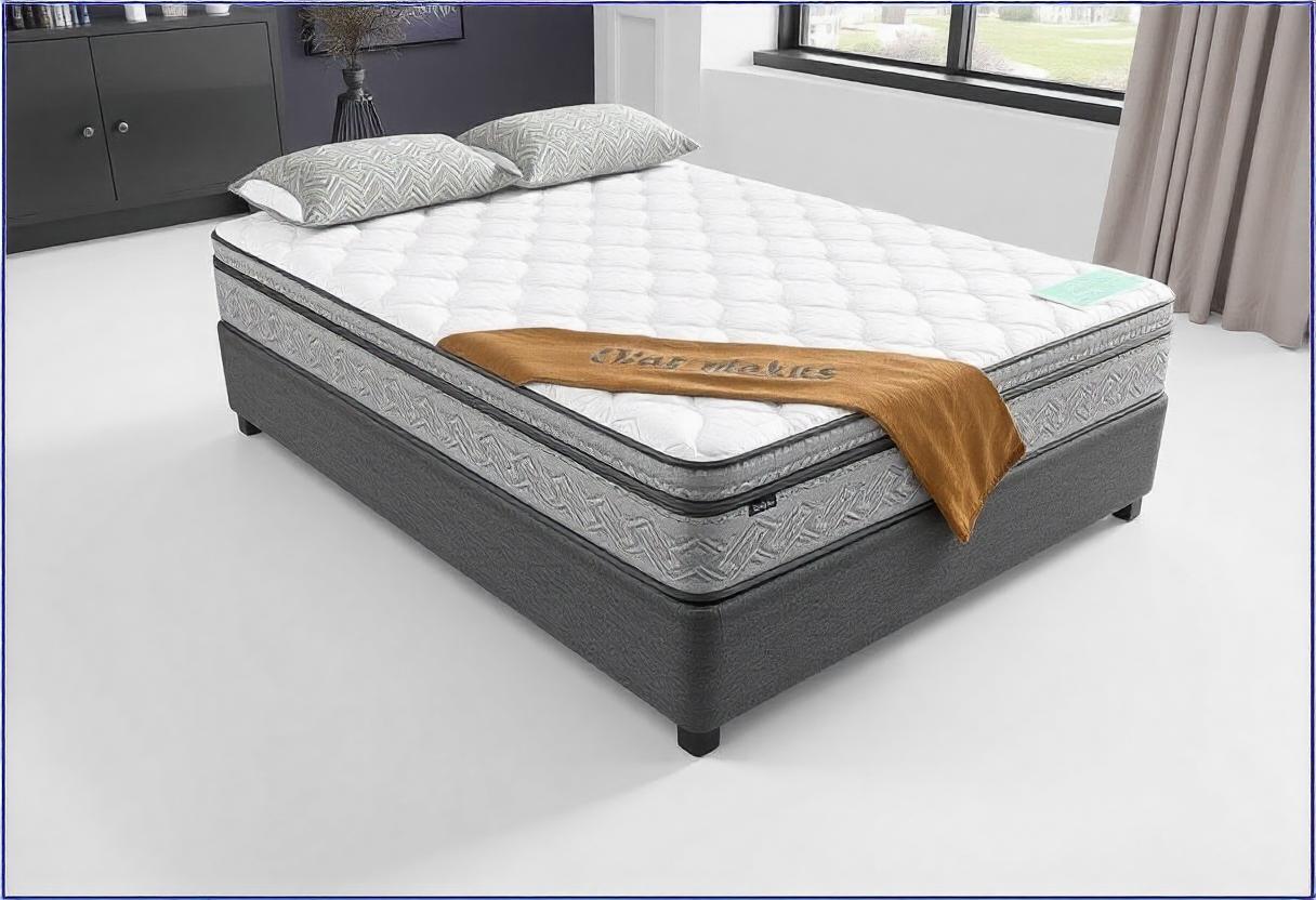 The photo shows a bed with a wooden frame. It is a king size mattress with indoor furniture such as pillows, bed sheets, duvet cover, and cushions. The design includes linens and a wall in the background.