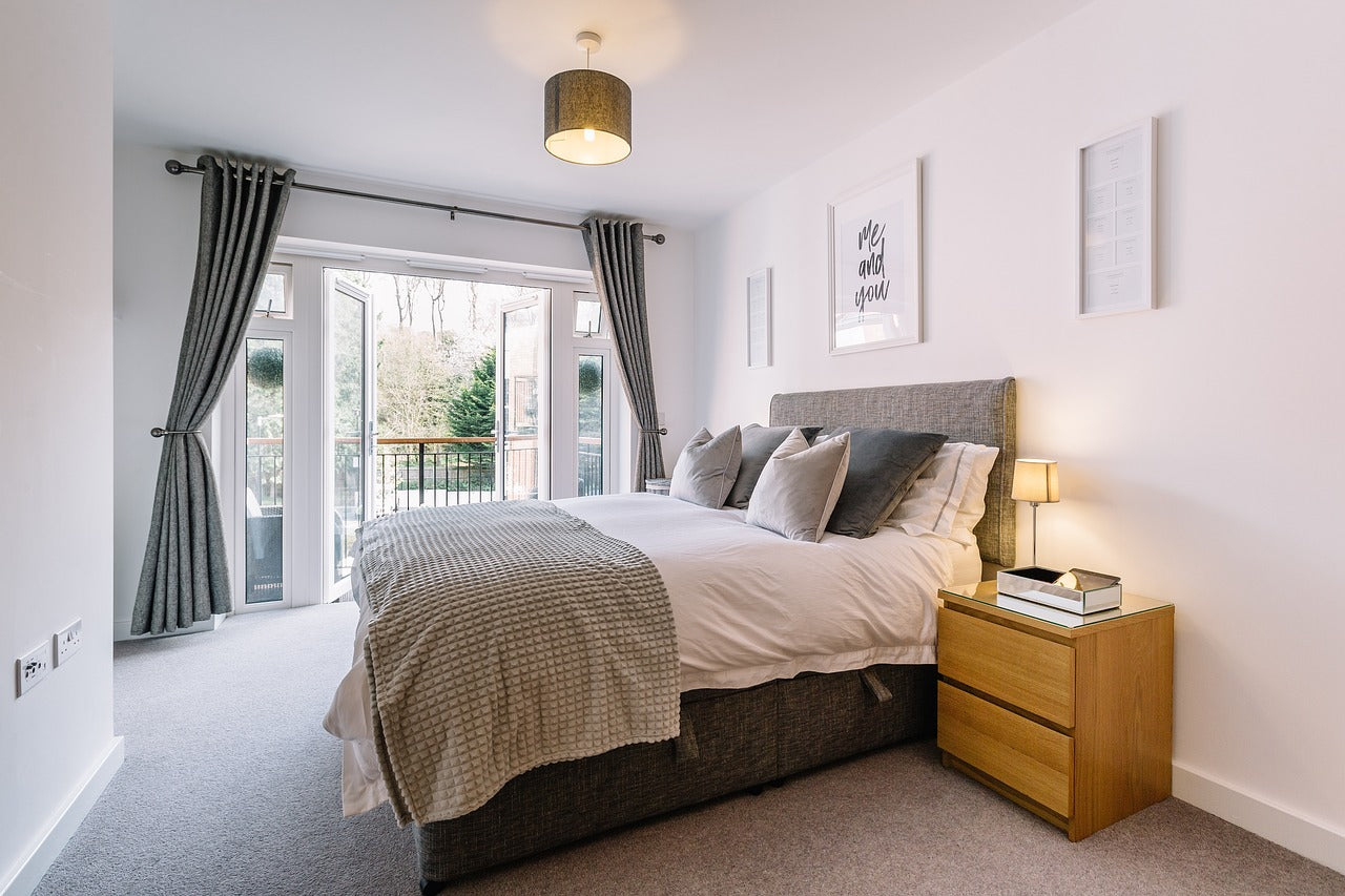 Bedroom with king size bed wooden bed frame, grey upholstered headboard, and wooden bedside table. Large glass doors with grey curtains open to a balcony.