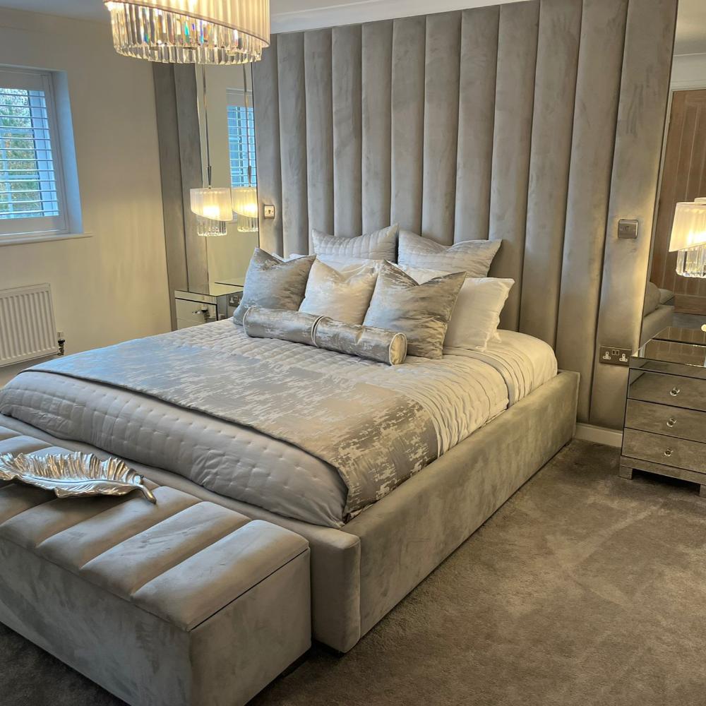 A luxurious bedroom with a gray ottoman single bed, matching pillows, and a large headboard, featuring mirrored side tables and pendant lights.