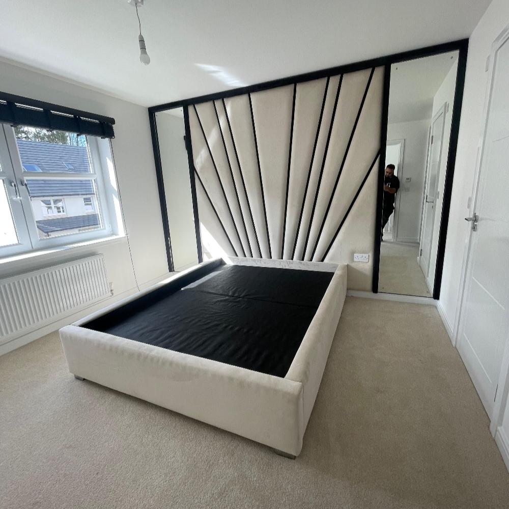 The image features the Freya Wall Panel in a modern minimalist bedroom. A light beige upholstered bed frame without a mattress is set against a striking accent wall with a fan-like geometric pattern. A large mirror reflects the room and a person capturing the photo. A window with a black blind allows natural light in, and a white radiator is positioned below. The neutral cream walls and carpet enhance the understated aesthetic.