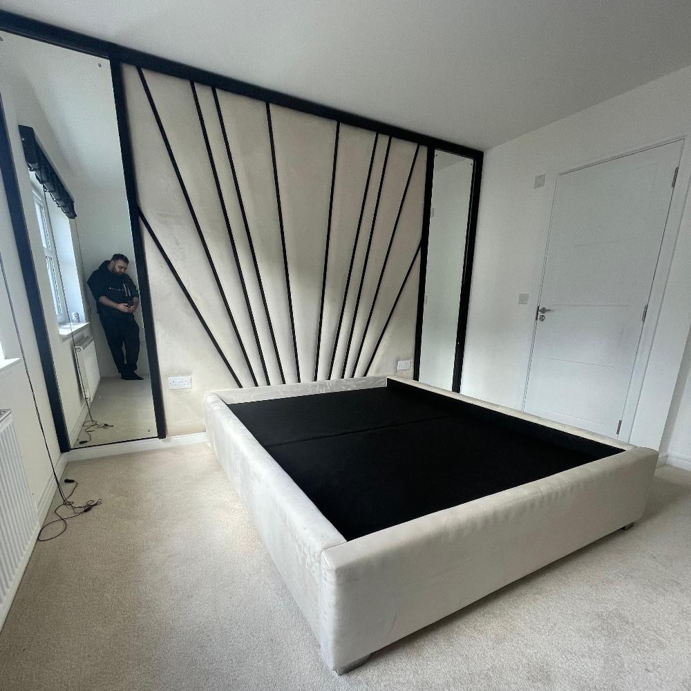 The image showcases the Freya Wall Panel in a modern bedroom featuring a large beige bed frame without a mattress. The striking headboard boasts a sunburst design made of large, fan-like cream panels with bold black lines. The bed sits atop an off-white carpeted floor. A large mirror on the left wall and another within the headboard's panels enhance the space, while natural light filters in through a partially visible window with a Roman blind. A man in dark clothing is reflected in the left mirror.