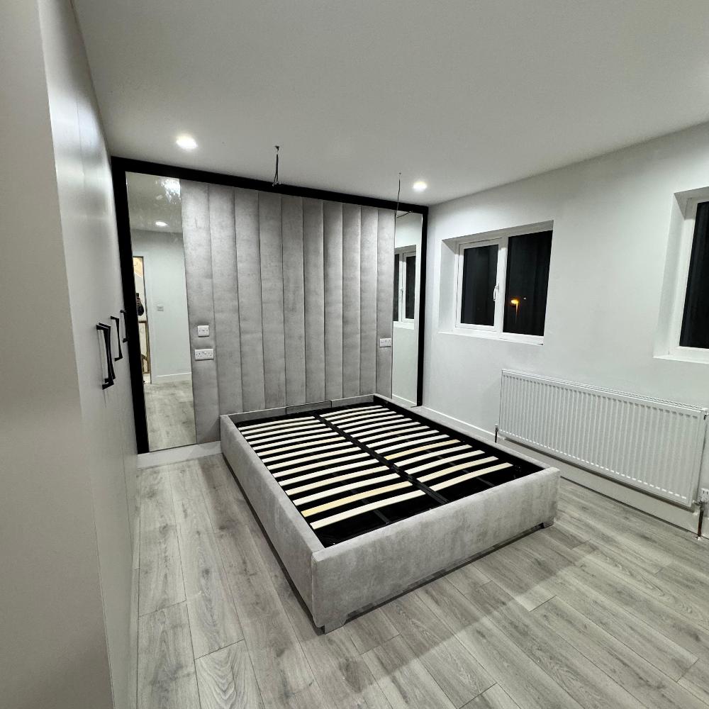 Minimalist bedroom with a gray luxe wall panel bed frame and vertical padded headboard, wooden floor, windows, and closet.

