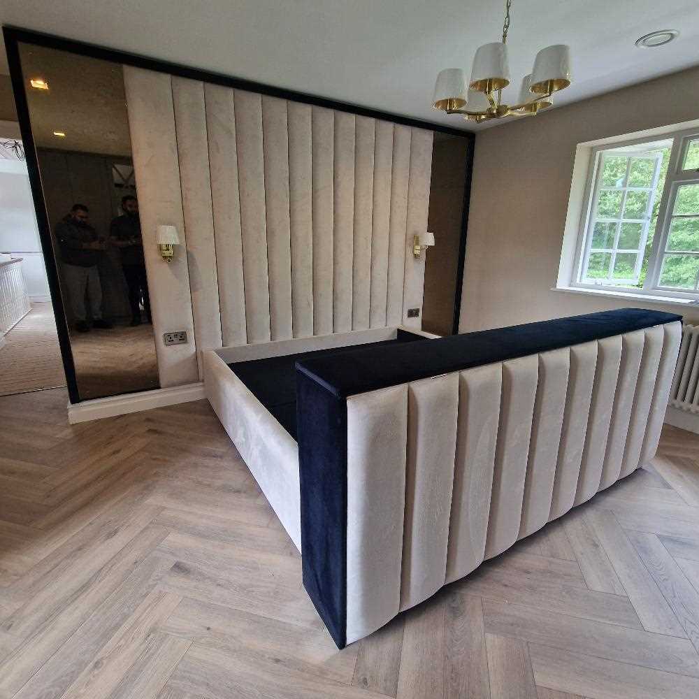 The image shows a modern, minimalist bedroom featuring a large wall panel bed frame with a design of vertical white padded panels bordered by black trim, situated against a matching padded wall. The floor is covered with light beige carpet, and the room is well-lit by natural light coming through two rectangular windows in the corner. The walls are painted in a light color, complementing the overall neutral color scheme of the room. 