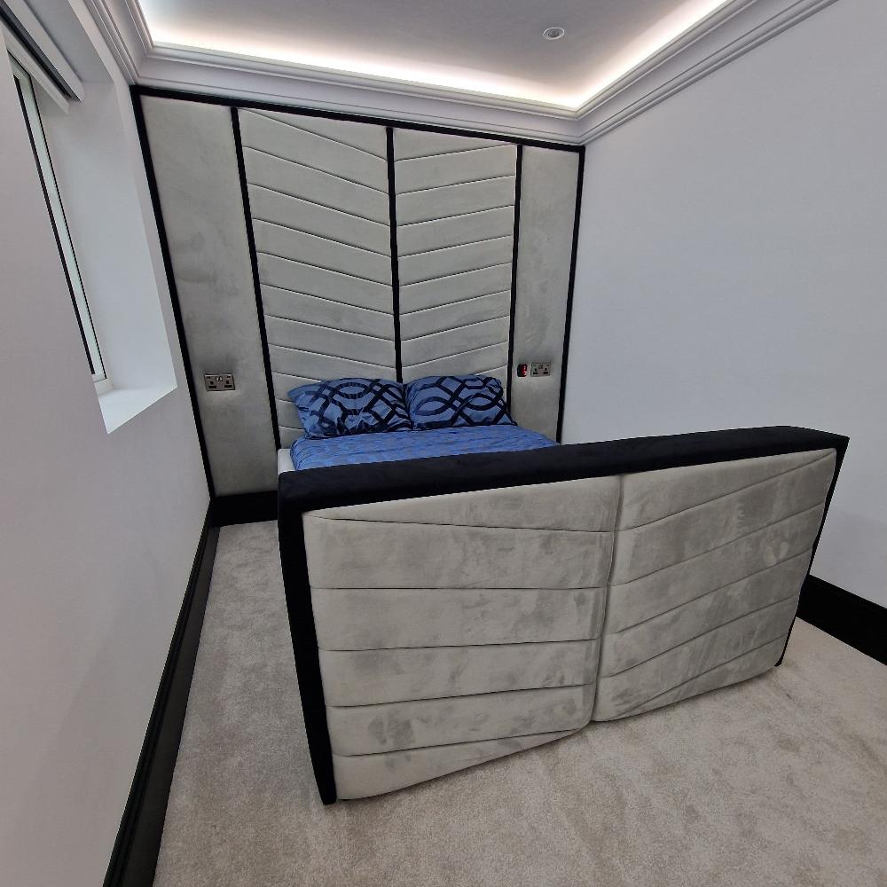 The image depicts a modern bedroom with a focus on a stylish Mr Herring Bone Design bed and headboard. The room features a sleek, minimalistic design with neutral tones. The bed is positioned against a large, upholstered headboard that covers much of the wall, composed of three panels with a chevron pattern. The central panel has diagonal stripes, creating a distinctive V-shape. The bed itself is made with a dark blue duvet and patterned pillows placed against the headboard. 