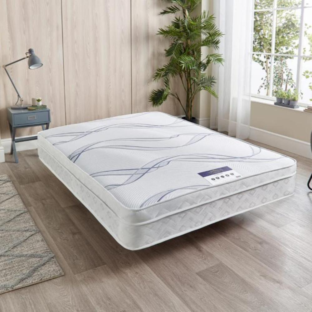 A white single pillow top 3000 pocket   mattress with gray and blue patterns on a wooden floor, next to a nightstand and a window with sheer curtains.

