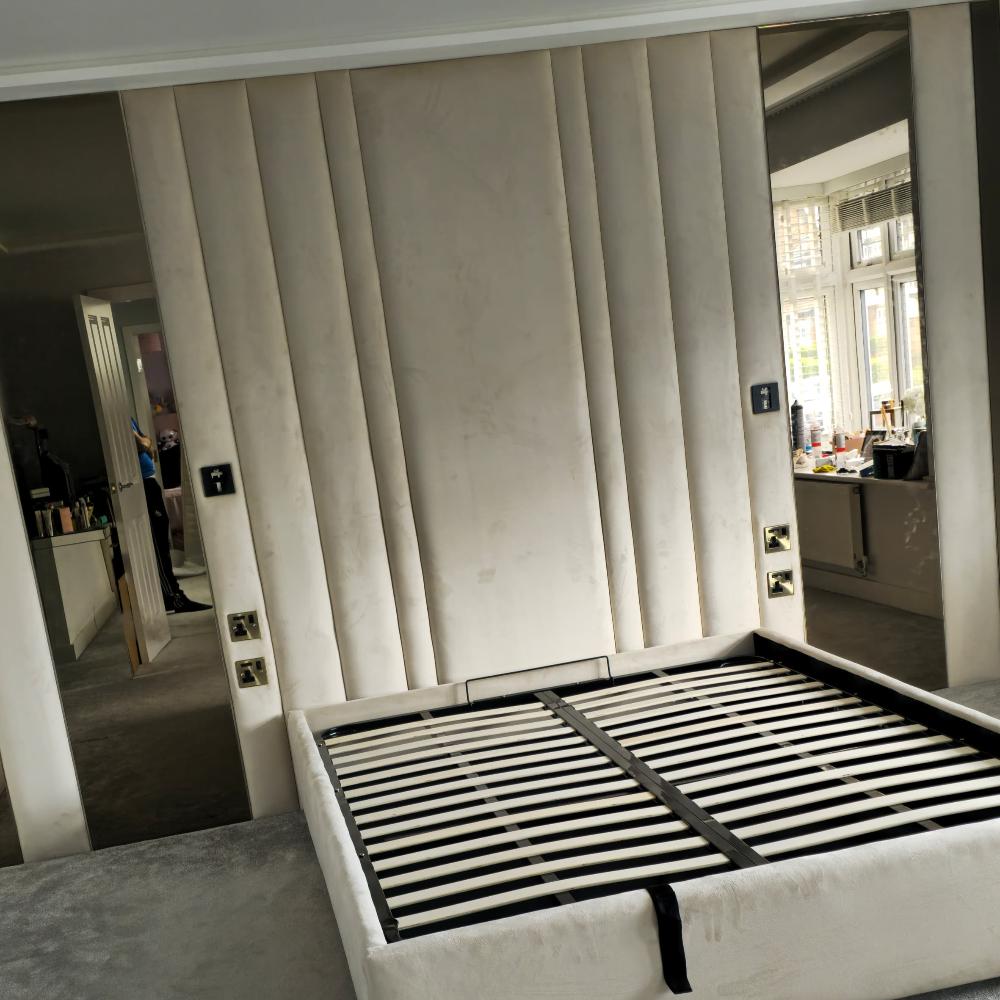 The image depicts a bedroom interior featuring a bed frame without a mattress. The alexander bronze panel bed frame has a cream fabric upholstered base with visible wooden slats. The backdrop consists of vertical panels with a suede-like texture in a light beige color. On either side of the panels, there are floor-to-ceiling mirrors. The room is carpeted in a light gray shade. To the right, a window with horizontal blinds allows natural light to enter, illuminating various items on a dresser or desk below.