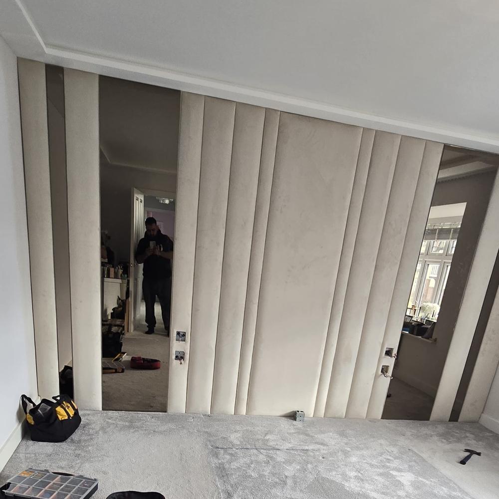 The image shows a room with a wall featuring vertical padded beige alexander bronze panels bed frame alternating with tall mirrors. The ceiling and walls are painted white, and the floor is covered with a light gray carpet. On the floor, there's a black and yellow tool bag and an open toolbox nearby. The mirrored sections reflect the image of a person standing in a doorway, taking the picture, and another part of the room with a window. 