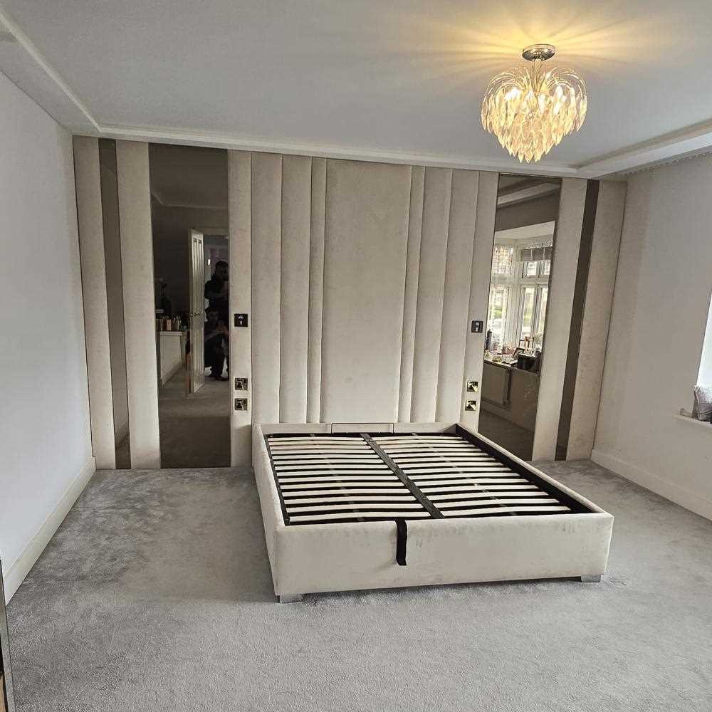 The image shows a modern bedroom with a panel bed frame positioned centrally on a soft, gray carpeted floor. The bed frame is upholstered in light beige fabric, and it has a slatted base, suggesting it is intended for use with a mattress. The wall behind the bed features vertical panels that match the bed's upholstery, flanked by two large mirrors. A window on the right side allows natural light to enter the room. The ceiling is white, with a decorative, leaf-like chandelier that emits a warm glow. 