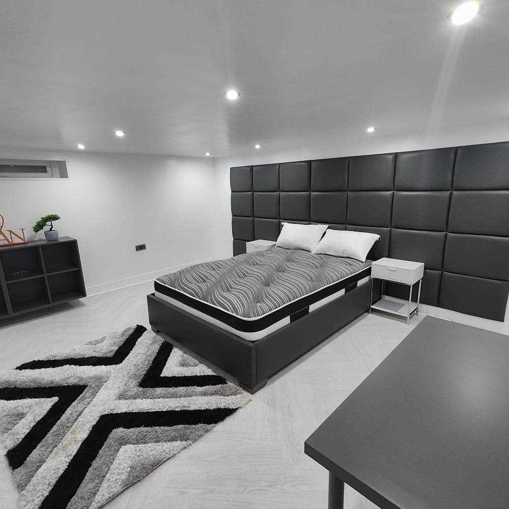 The image depicts a modern and minimalistic bedroom. At the center is a bed with a simple frame, dressed in a striped gray and black comforter and two white pillows. The wall behind the bed is covered with padded dark black panels, adding texture and depth. To the right of the bed, there is a sleek, square nightstand with a metal frame and a small shelf. The room features a light gray wooden floor, partially covered by a plush rug with a geometric pattern in shades of black, white, and gray. 