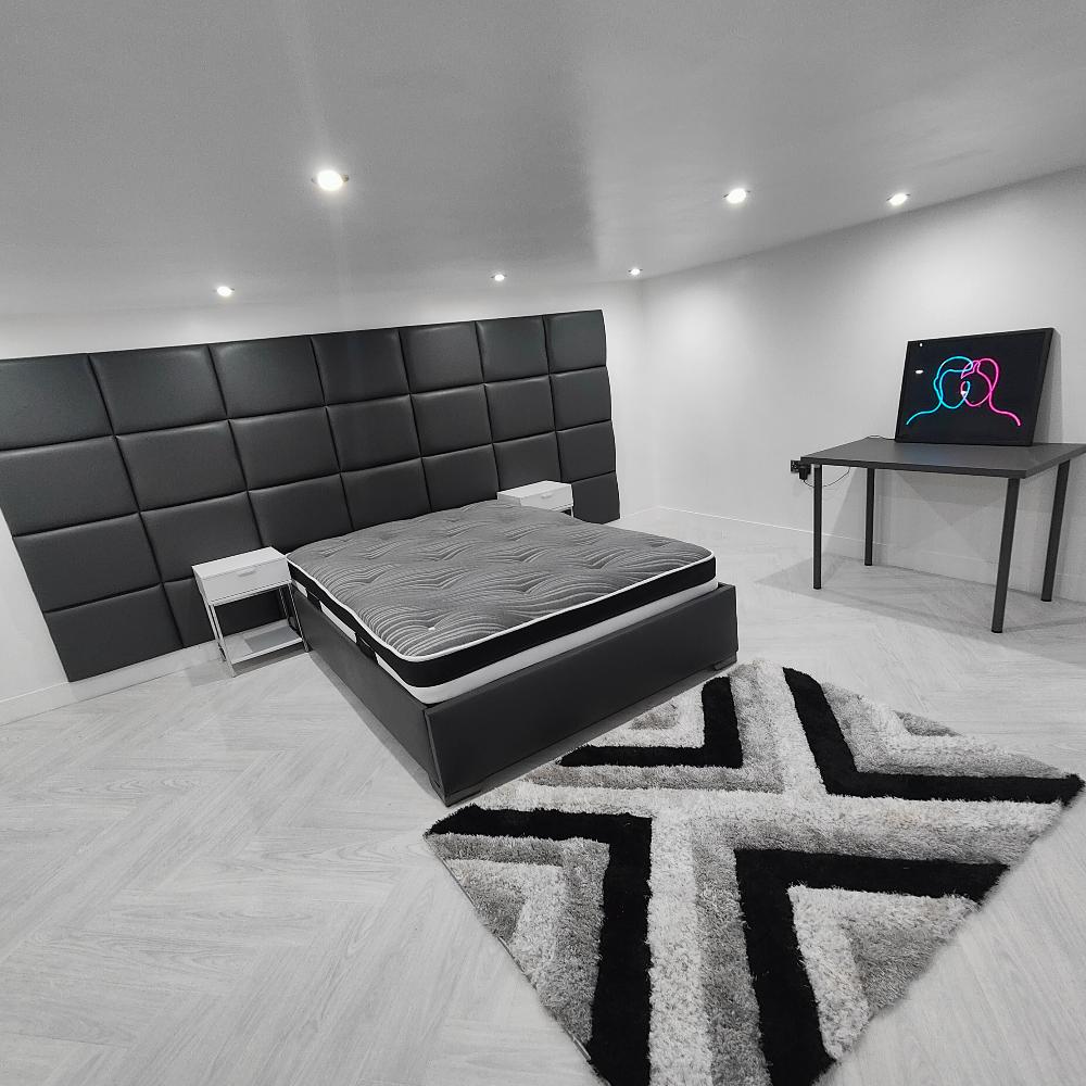 The image shows a modern bedroom with a minimalist design. The room features a large black wall panel bed with a  padded headboard made up of square panels, creating a contemporary look. The bed has a gray mattress and is positioned centrally against the wall. On either side of the bed are small, white bedside tables. The floor is covered with light wood-patterned tiles, and there is a plush area rug in front of the bed. 