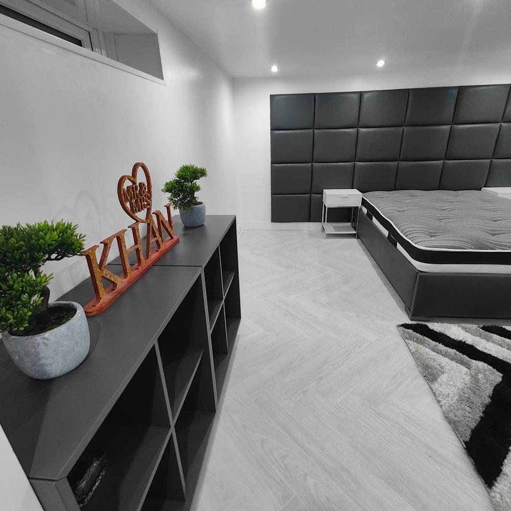 The image depicts a modern bedroom with a minimalist design. On the left side, there is a dark gray shelving unit with six open compartments. On top of this unit are two small green potted plants. Between the plants is a decorative wooden sign featuring the text “MR & MRS KHAN.” The bedroom floor is covered with light-colored wooden flooring arranged in a herringbone pattern. On the right side of the image, a black wall panel bed with a gray upholstered headboard and mattress is visible.