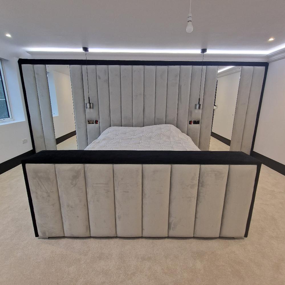 The image shows a modern bedroom with a large bed as its focal point. The Dantel Bespoke bed is integrated into a rectangular frame that surrounds it on three sides, forming a canopy-like structure. The frame and bed are upholstered in a soft, light gray fabric with vertical tufting, contrasted by black edges. The headboard and sides are tall, giving the room a luxurious feel. The bedspread is white with subtle geometric patterns.  