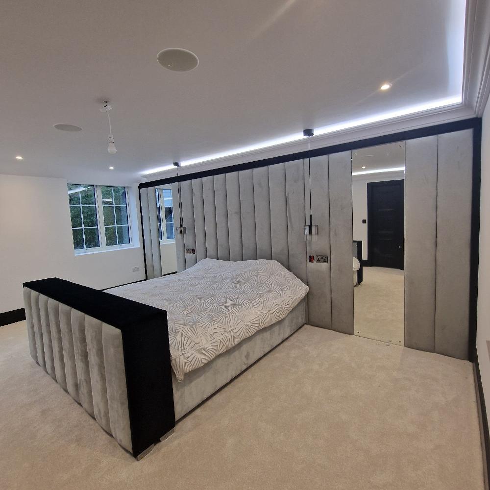 The image showcases a modern minimalist dante bespoke panel bedroom. The room’s color palette includes soft grays, blacks, and whites. Dominating the foreground is a plush bed with a padded headboard and footboard featuring vertical panels in light gray fabric with black trim. The bed is covered with a white geometric patterned duvet. The back wall is lined with matching light gray padded panels, creating a cohesive design. Suspended above the bedside are two small, cylindrical pendant lights. 