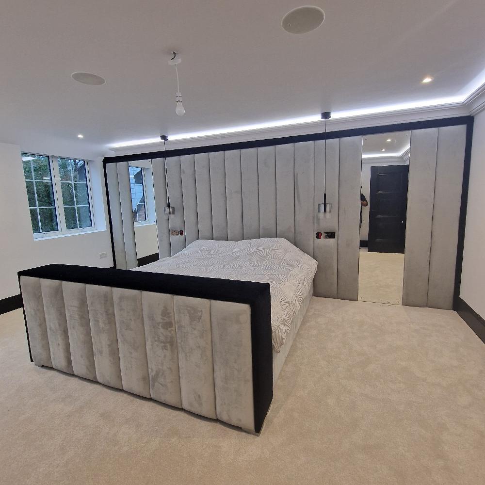 The image shows a modern, minimalist bedroom with a focus on symmetry and clean lines. At the center is a large bed with an upholstered headboard and footboard, both featuring vertical, padded grey panels bordered with black. The Dantel Bespoke Panel bed is covered with a patterned white duvet. The wall behind the bed is mirrored, with the mirrored sections also framed by identical padded grey panels, enhancing the room's spacious appearance. Above, a simple bulb hangs from the ceiling.