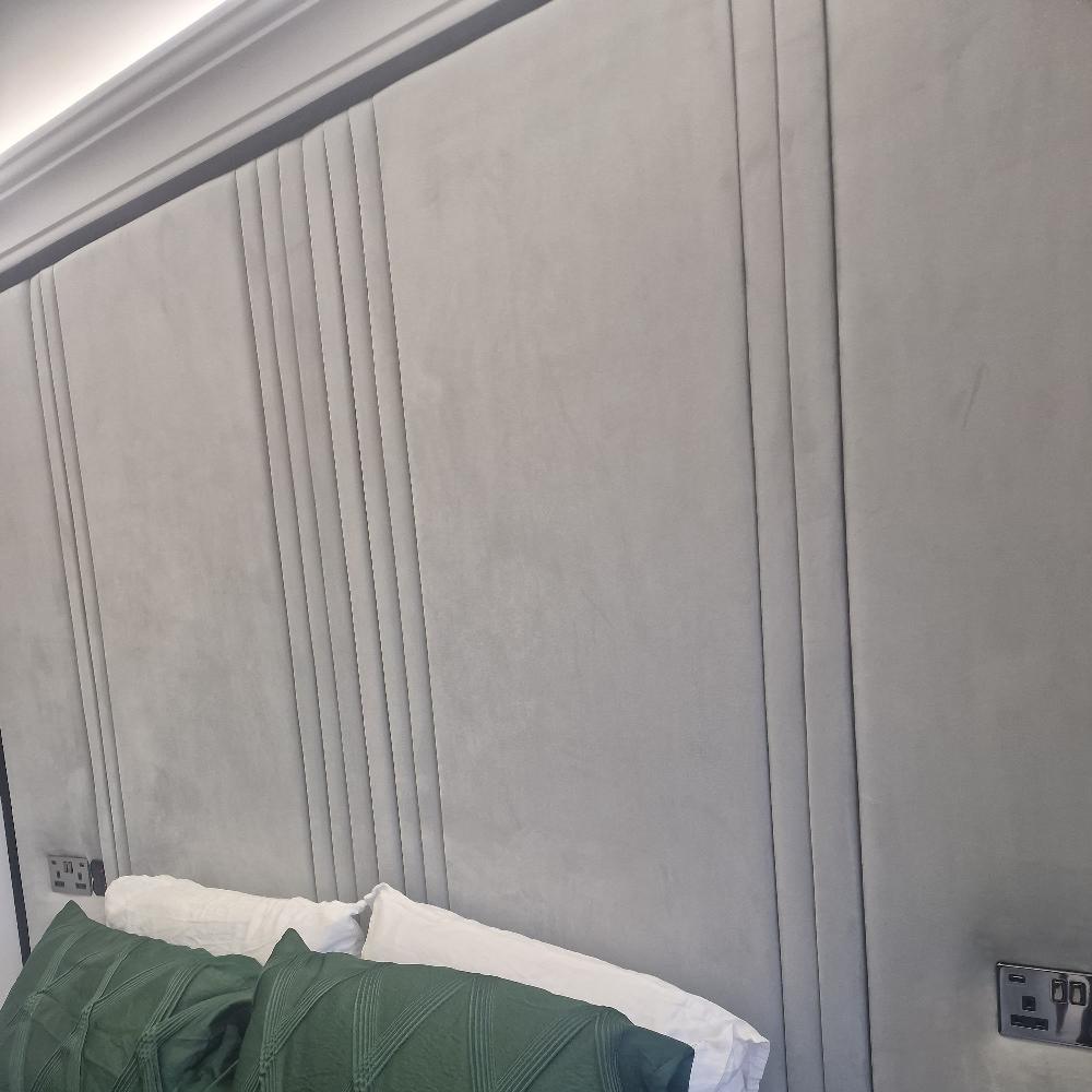 The image showcases The Eleanor Panel Design with light gray fabric panels featuring vertical stitched details and evenly spaced seams for a textured, modern look. Below, the bed is styled with white and dark green pillows, flanked by wall-mounted electrical sockets on either side for added convenience.