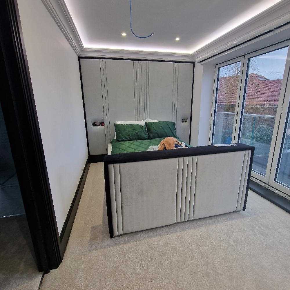 The image showcases a modern bedroom centered around The Eleanor Panel Design, featuring a tall light gray upholstered headboard with vertical grooves that match the footboard. The bed is styled with moss green bedding and beige accents, adding warmth to the soft neutral space. Light carpet flooring and dark trim enhance the sophisticated look, while a sliding glass door on the right opens to a terrace view. Recessed and cove lighting provide a bright, welcoming ambiance.