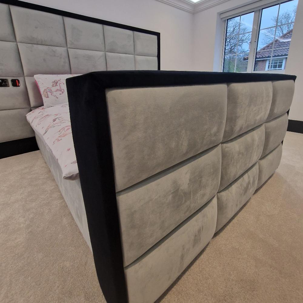 The image showcases The Manhattan Design bedroom featuring a plush upholstered bed with a tall, grid-patterned gray headboard and footboard framed in black. The bedding displays a subtle pink unicorn and star pattern. Neutral-toned carpet lines the floor, while a large window reveals a view of a tiled roof and bare trees outside, allowing natural light to fill the room.