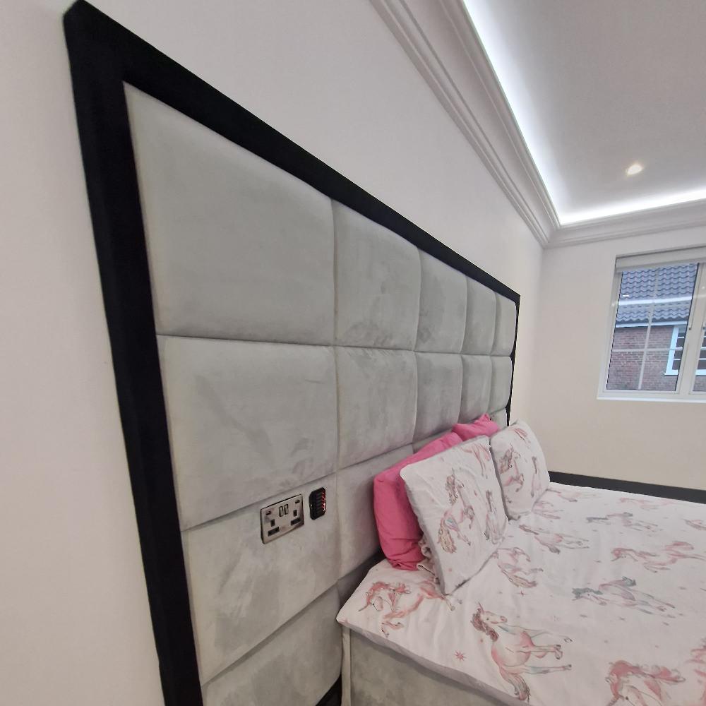 The image features The Manhattan Design bedroom with a large gray upholstered headboard framed in black. It includes a dual electrical outlet and a digital display. The bed is adorned with white unicorn-patterned bedding and pink pillows. A window with white blinds lets in light, showcasing a tiled roof outside. Modern recessed strip lighting provides ambient illumination.