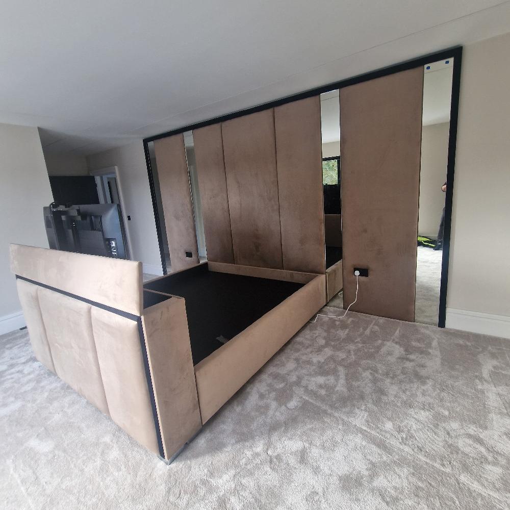 The Samsun Wall Panel TV Bed features a minimalist light brown upholstered frame on plush carpet. Tall panels behind the bed include mirrored sliding doors for the wardrobe, reflecting the room. Reflective strips enhance the contemporary aesthetic, complemented by a wall-mounted television and neutral light-colored walls.