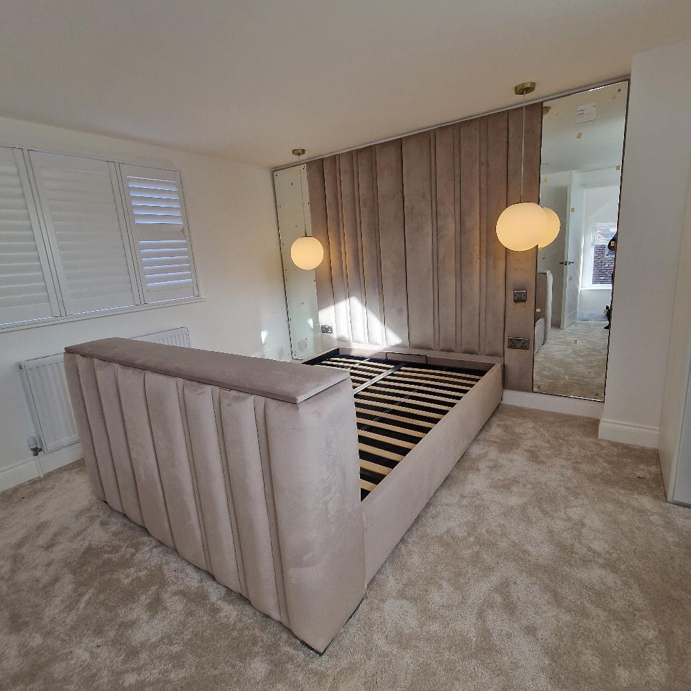 The image depicts a modern bedroom with a bed frame and stylish decor. The bed frame is upholstered in a soft beige fabric with vertical tufting, matching the plush carpeting. Behind the bed is a padded accent wall with vertical panels, flanked by large mirrored surfaces on either side. Two spherical pendant lights hang from the ceiling in front of the mirrors, casting soft illumination. On the left side of the room, there are white plantation shutters covering a window, with a radiator positioned below.