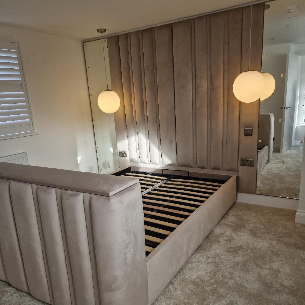 The image shows a modern, well-lit bedroom with a beige color palette. A large, upholstered beige split panel bed frame with vertical stitching and no mattress dominates the room. It sits on a plush, light beige carpet. Behind the bed is a mirrored wall, and a paneled headboard runs vertically in a matching beige fabric. Two spherical pendant lights hang from the ceiling in front of the mirror, providing ambient light. On the left, there are white shutters covering a window with sunlight filtering through.