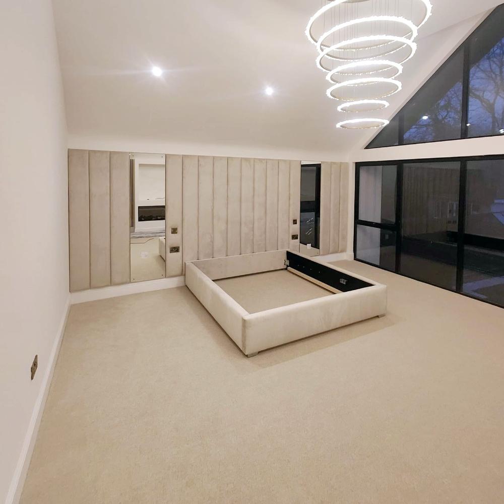 The image depicts a modern, minimally furnished bedroom with a high, sloping ceiling. The room features a large, light-colored, carpeted floor. In the center, there is a platform amaya panel bed frame without a mattress. The walls have vertical padded panels in a soft beige fabric, giving the room a luxurious feel. Two rectangular mirrors are embedded within the paneling, one on each side of the wall. 