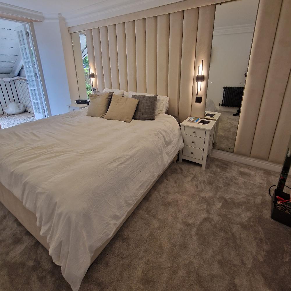 The image shows a modern, well-decorated bedroom. The centerpiece is a large amaya panel bed with a white duvet and three large pillows in varying shades of beige and gray. The bed is positioned against a plush, vertically paneled headboard matching the color of the pillows. On either side of the bed, a narrow mirrored wall features contemporary vertical light fixtures. To the right side of the bed is a small, white nightstand with two drawers. 