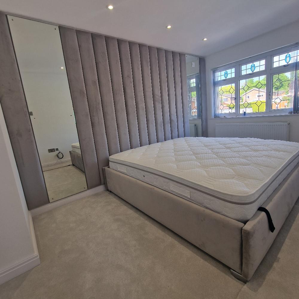 The image portrays a modern, minimalist bedroom interior featuring a large bed with a padded headboard. Amaya panel bed is covered with a white quilted mattress and is set against a wall with vertical, upholstered panels in a muted grey color. The upholstery creates a textured backdrop running from floor to the ceiling. To the left, there is a floor-to-ceiling mirror which reflects part of the room. Natural light enters through a trio of leaded glass windows adorned with blue and yellow geometric designs.