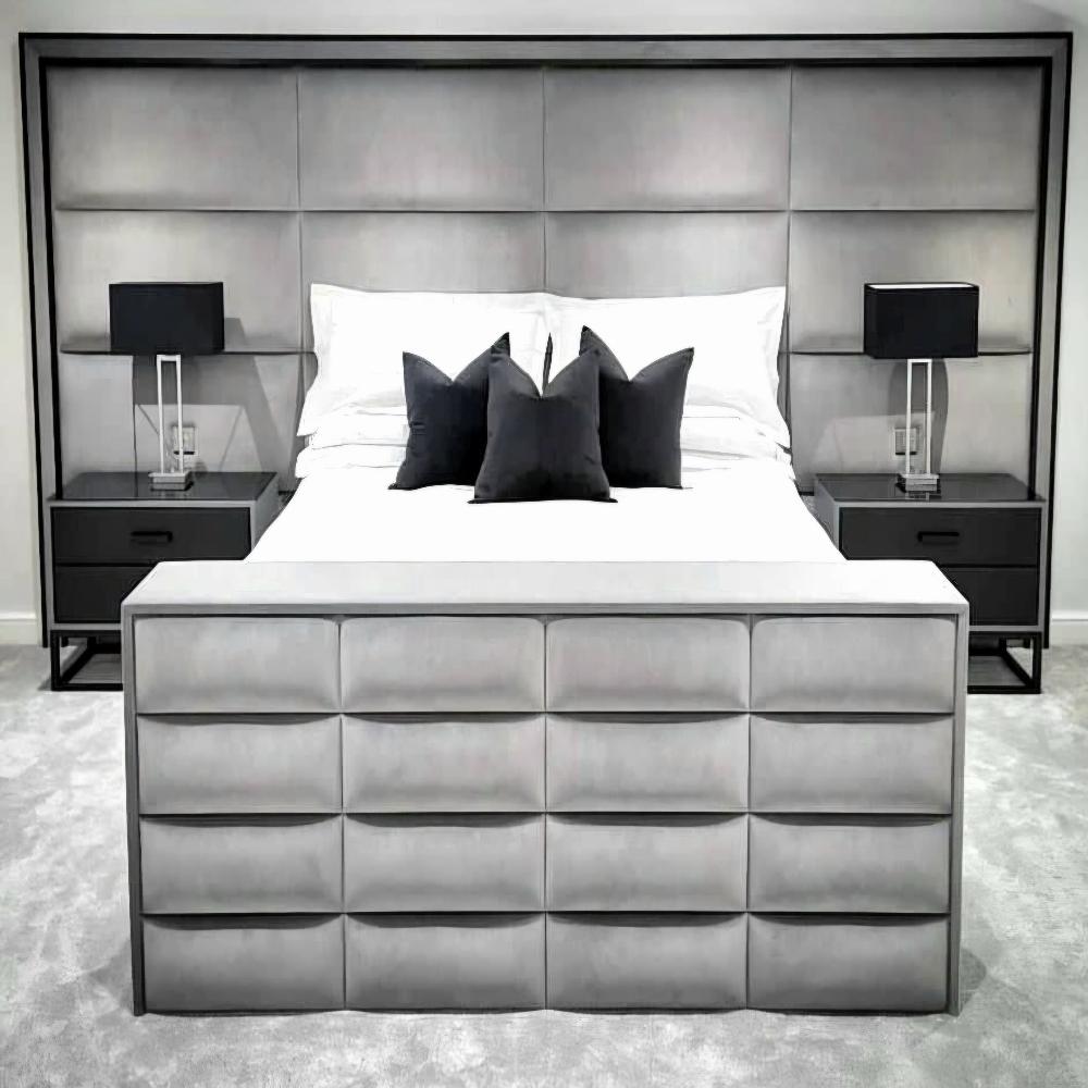 The image shows a modern and stylish bedroom setting. The central focus is a large aries panel bed with a gray upholstered headboard that extends across the width of the wall. The bed is made with crisp white sheets and is adorned with three black decorative pillows in front of the headrest. Flanking the bed on either side are two matching nightstands with dark finishes. Each nightstand holds a square black lamp with a geometric base. The foot of the bed features a padded bench with similar gray upholstery.