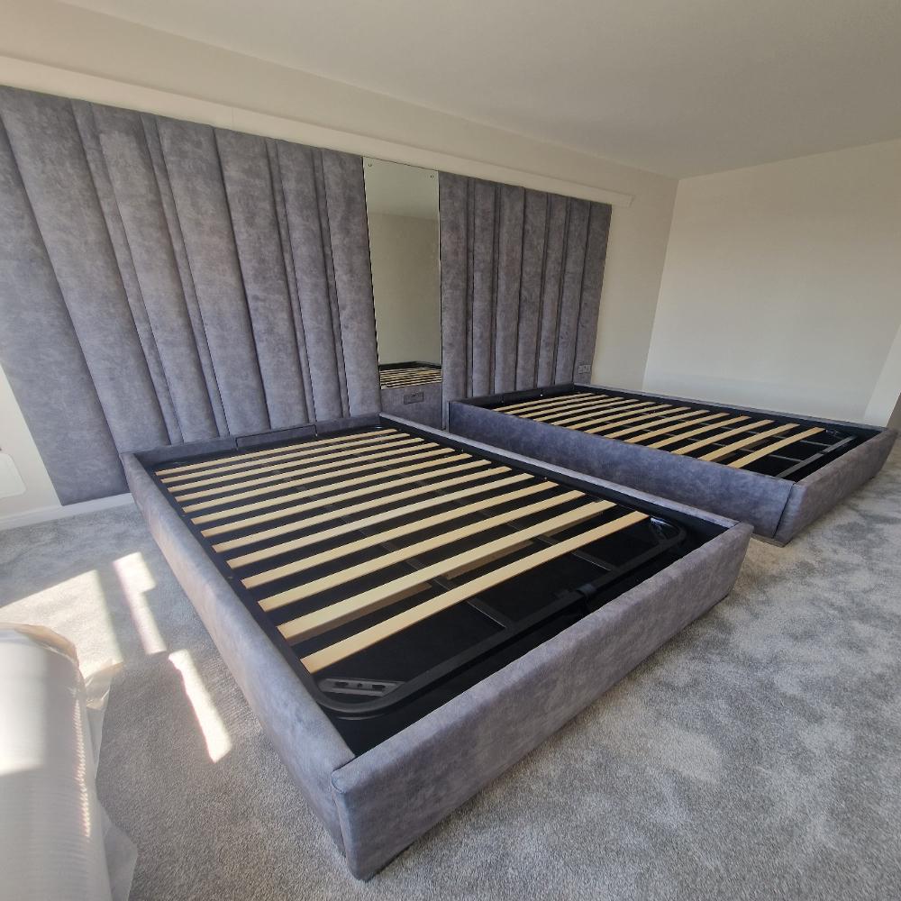 Two grey upholstered duo panel bed frames with wooden slats in a bedroom with a padded wall and a large mirror.