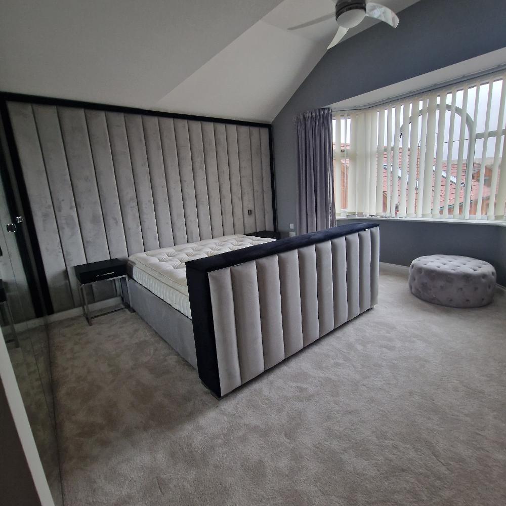 The image showcases a modern, elegant bedroom featuring The Elise TV Panel Bed. The large bed boasts a gray upholstered headboard and footboard with vertical paneling that complements the full wall behind it. Covered with a light mattress, the bed is paired with a sleek black side table with a glass top on the left. A plush, light gray carpet covers the floor, while a large window with vertical white blinds on the right lets natural light into the room, accented by a gray curtain. 