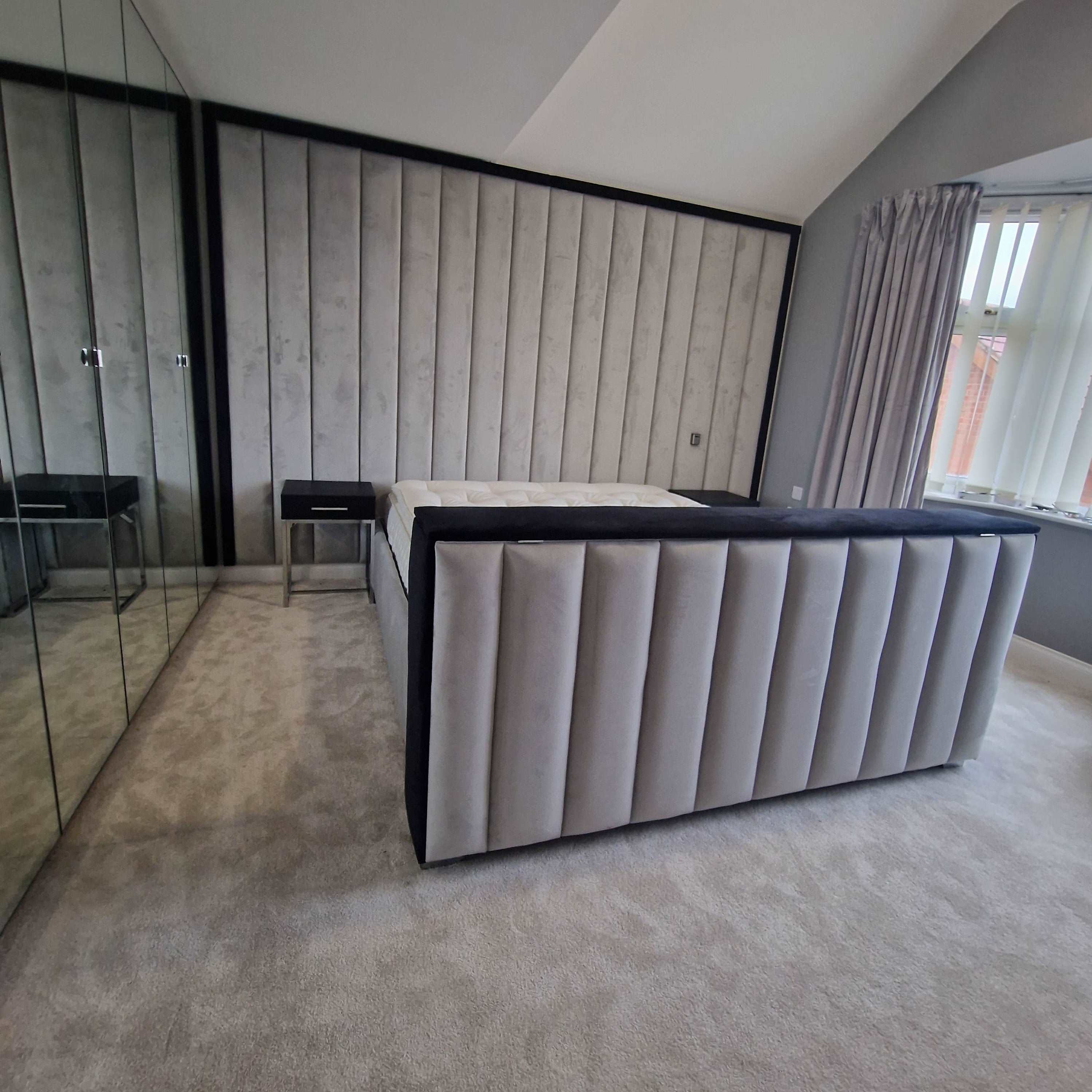 The image depicts a modern bedroom featuring The Elise TV Panel Bed at its center. This large, cushioned bed boasts a high, vertically-panelled headboard and footboard, upholstered in soft gray fabric with dark trim. A plush, light gray carpet covers the floor. To the left, a mirrored wardrobe stretches along the wall, reflecting parts of the room. Next to the bed, a sleek black nightstand with a single drawer and metal legs adds a touch of elegance. 