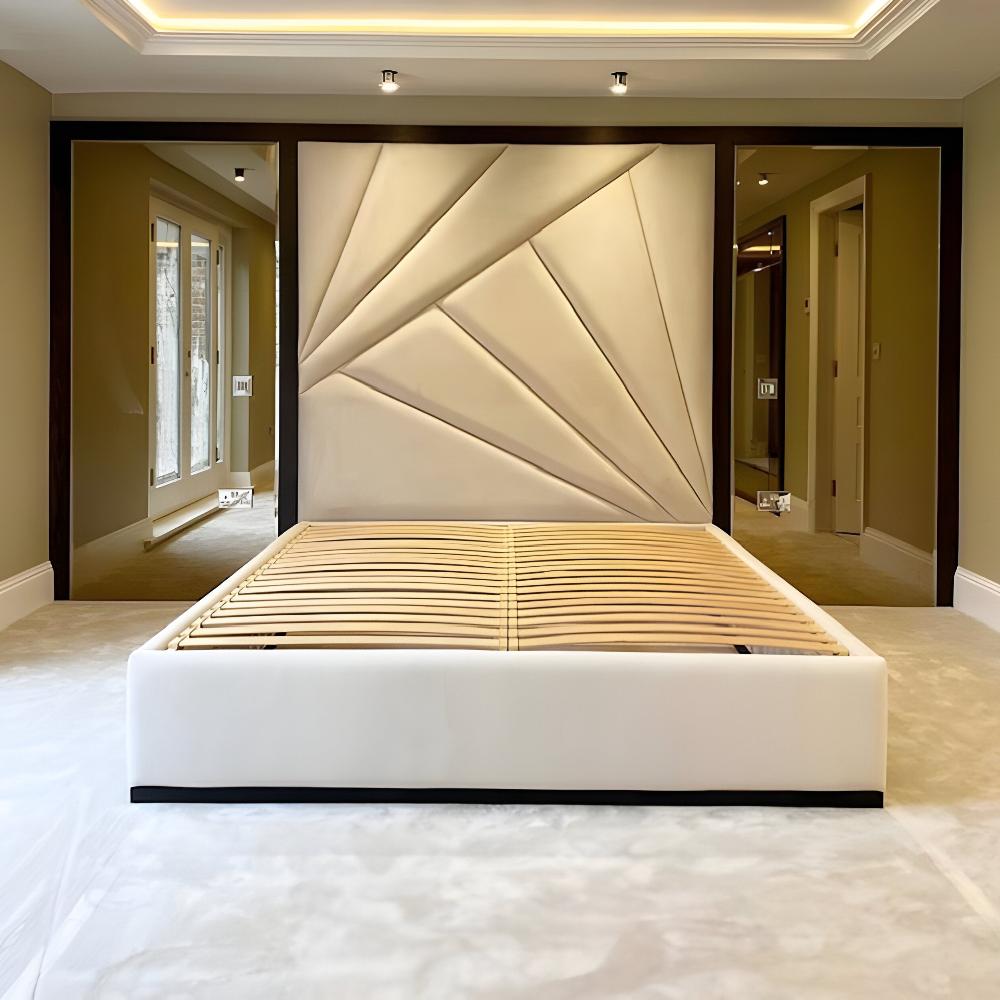 The image features The Linear Panel Bed Frame with Bronze Mirrors in a contemporary bedroom. The stylish bed has a unique headboard of large, geometric panels in smooth beige fabric, framed by dark wood. Its white upholstered base reveals wooden slats beneath the mattress. The room boasts a high ceiling with recessed lighting and soft neutral walls. Large glass doors on the left allow natural light, while a doorway on the right adds depth. The carpeted floor enhances the serene atmosphere.