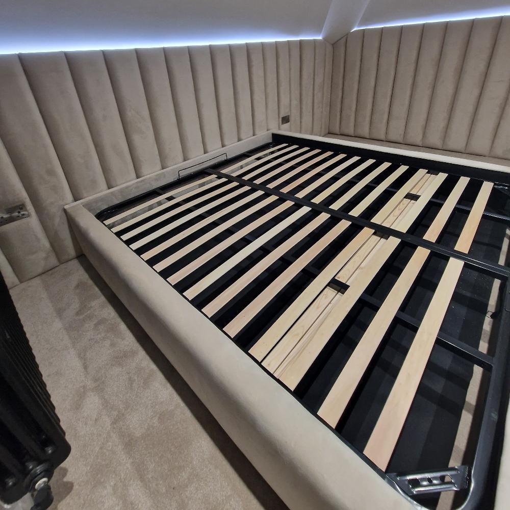 The image showcases The Loftly Panel Bed Frame featuring an upholstered headboard and side panels in plush beige fabric. The bed base is supported by black metal with wooden slats across the width. Surrounding walls are covered in the same beige fabric, creating a textured vertical panel pattern. The bed sits on a light beige carpet, with a sleek black radiator visible on the left. A strip of LED lighting at the top of the headboard provides ambient illumination against the wall.

