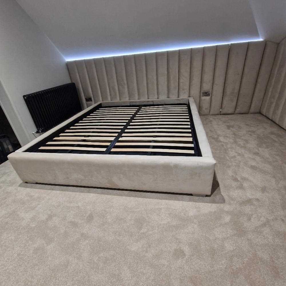 The image showcases The Loftly Panel Bed Frame in a minimalist room with a sloped ceiling. The cream upholstered platform bed frame features visible wooden slats, while the headboard and side walls are padded with vertically lined cream panels. The matching carpet maintains the monochromatic theme, and a black radiator on the left provides contrast. Soft lighting glows from under the sloped ceiling, enhancing the modern atmosphere.