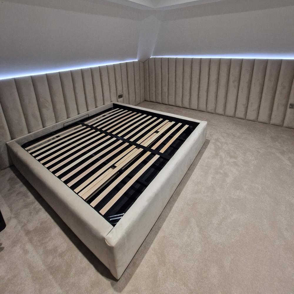 The image features The Loftly Panel Bed Frame in a minimalist bedroom. The large bed frame, upholstered in light beige fabric, is centered and low-profile, with visible wooden slats for mattress support. The room boasts wraparound quilted wall paneling in a matching beige, enhancing texture and cohesion. The carpeted floor complements the neutral tones, while subtle LED lighting along the top of the paneling casts a soft glow throughout the space.


