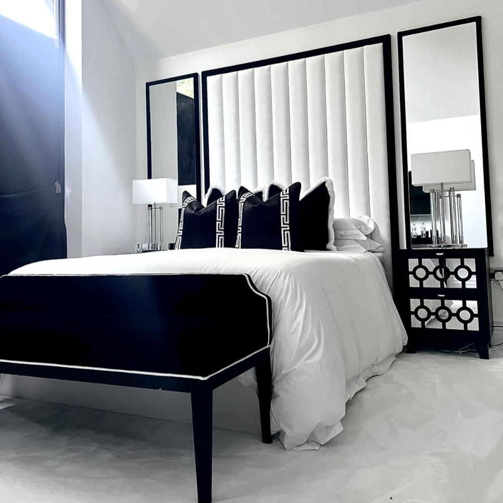 The Mont Blanc Bed Frame takes center stage in a chic, monochrome bedroom. The white vertically tufted headboard, framed in black, is paired with mirrored panels and matching black bedside tables. Plush black pillows, a pristine white comforter, and a black bench complete the sophisticated look.