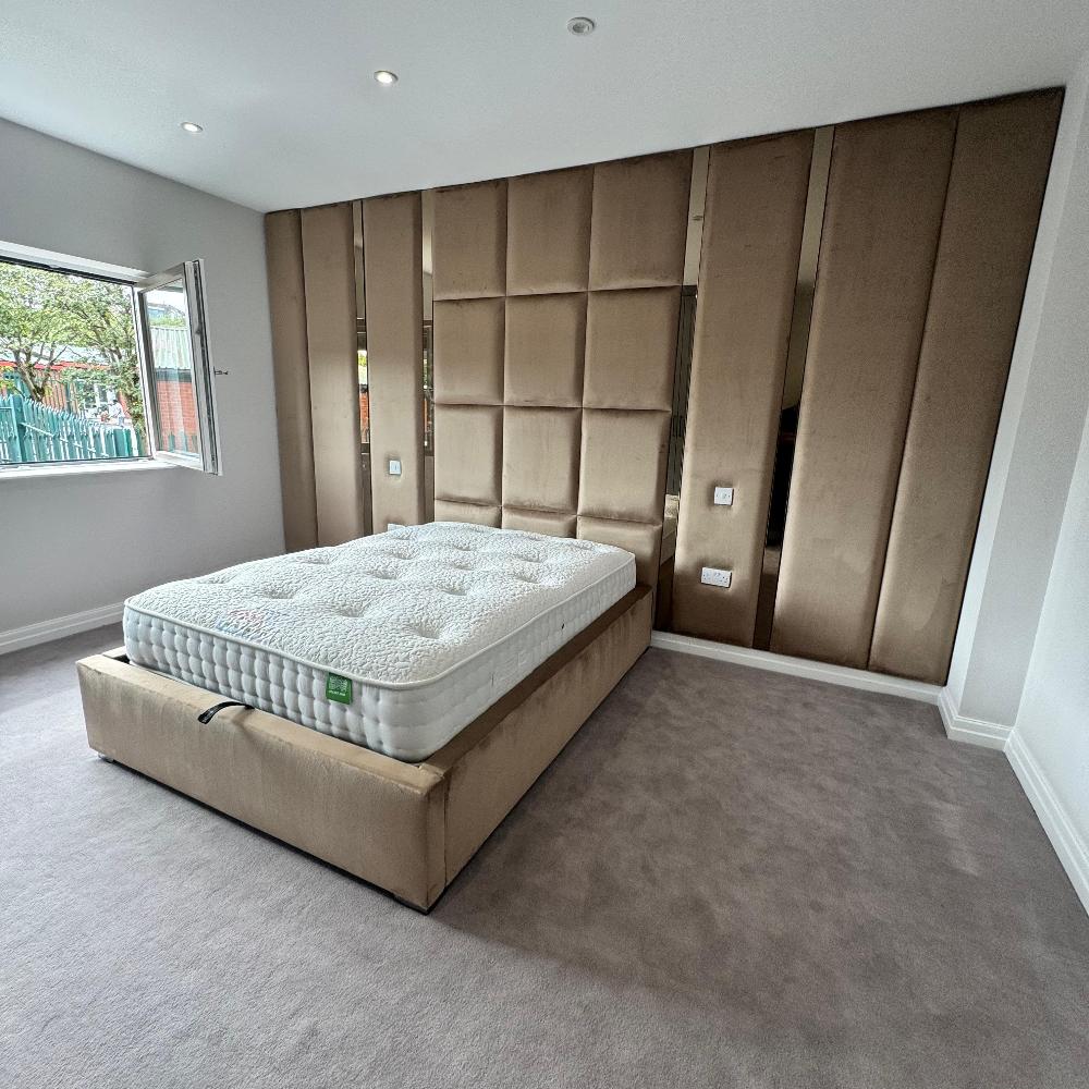 The Nexus Hybrid Wall Panel creates a cohesive look in this minimalist bedroom with beige vertical and square upholstered panels. The matching padded bed frame and neutral tones blend seamlessly, while natural light from the open window highlights the room's simplicity and elegance.