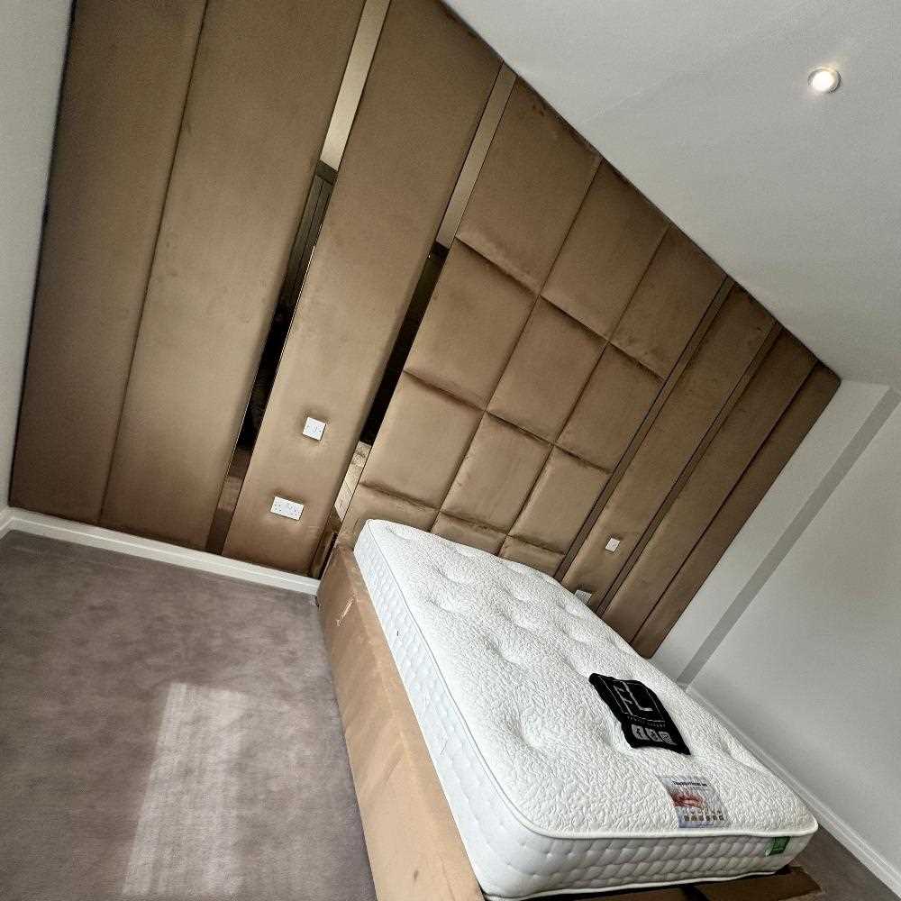 The Nexus Hybrid Wall Panel elevates this modern bedroom with its expansive plush headboard featuring vertical and square brown panels. A bare tufted mattress sits on a base wrapped in protective cardboard, while subtle lighting and neutral tones complete the contemporary look.