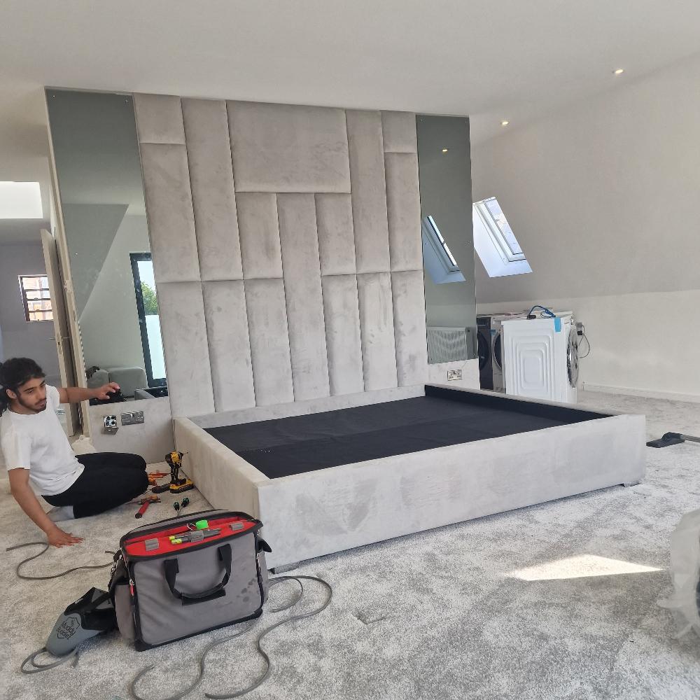 The Rowells Wall Panel Bed, upholstered in light gray fabric, is central to this modern bedroom under renovation. Its geometric padded headboard features vertical and horizontal panels. A person sits on the plush gray carpet, organizing tools, while a large floor-to-ceiling mirror reflects the spacious room. A white washing machine is temporarily positioned against the wall. Natural light floods in through skylights in the slanted ceiling, enhancing the minimalist decor with soft gray tones.