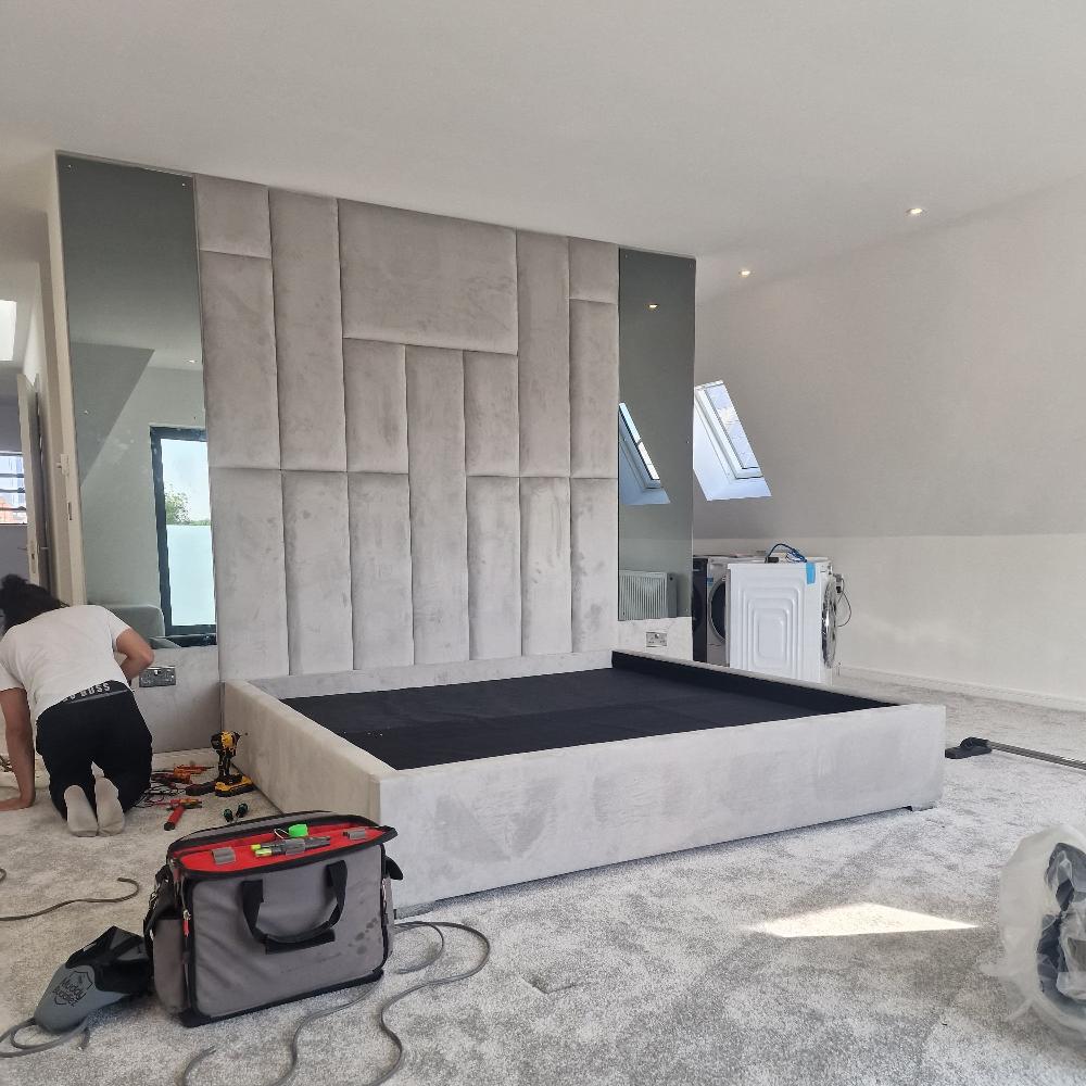 The Rowells Wall Panel Bed stands prominently in this modern bedroom setup, featuring a geometric textured headboard made of varying rectangular panels. Tall, narrow mirrors flanking the headboard enhance the room's sense of space. The light gray carpet and white walls create an airy ambiance, while a washing machine temporarily placed near the slanted ceiling adds a functional element. 