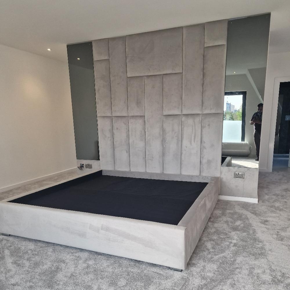 The Rowells Wall Panel Bed serves as the centerpiece of this minimalist bedroom, featuring a light gray upholstered frame and a textured headboard with vertically aligned rectangular panels. Flanking vertical mirror panels reflect the serene space, while plush gray carpeting and white walls enhance the clean aesthetic. A glimpse of greenery and a distant building appears in the right mirror's reflection, alongside a person near the doorway.