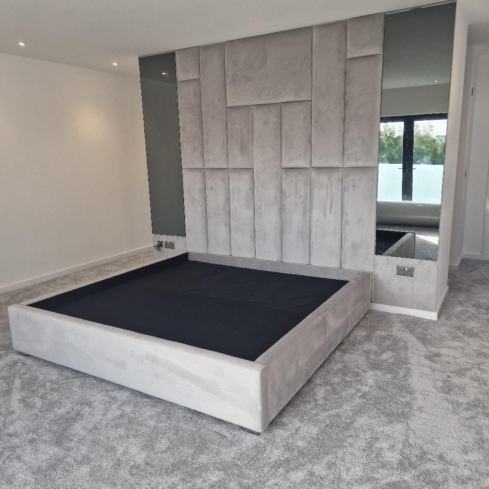 The Rowells Wall Panel Bed anchors this modern bedroom, featuring a large light gray upholstered frame. A geometric accent wall adds depth, flanked by tall floor-to-ceiling mirrors. The plush gray carpet and recessed ceiling lights enhance the monochrome theme, while a window reveals greenery outside.