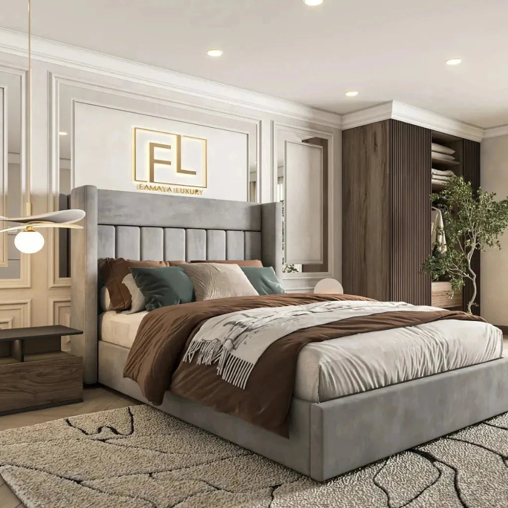 The Savoir Wing Back Bed Frame is featured in a luxurious bedroom with a tall, light gray upholstered headboard. The bed is styled with neutral bedding and decorative pillows. A wooden nightstand holds a geometric lamp. Elegant white walls with molding and a framed "FL" logo enhance the space. A built-in closet displays organized clothes, and a potted plant adds a touch of greenery. The room is completed with a plush light-colored carpet.