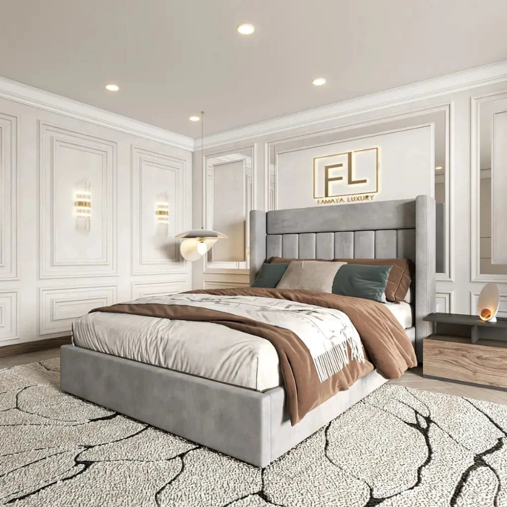 The image features a luxurious bedroom centered around a Savoir Wing Back Bed Frame with a light gray upholstered headboard. The bed is styled with white and brown bedding, complemented by neutral pillows. Flanking the bed are contemporary bedside tables with decorative lamps. The light cream paneled walls are illuminated with soft ambient lighting, and a monogram sign is mounted above the bed. A cream and black patterned rug enhances the sophisticated decor.