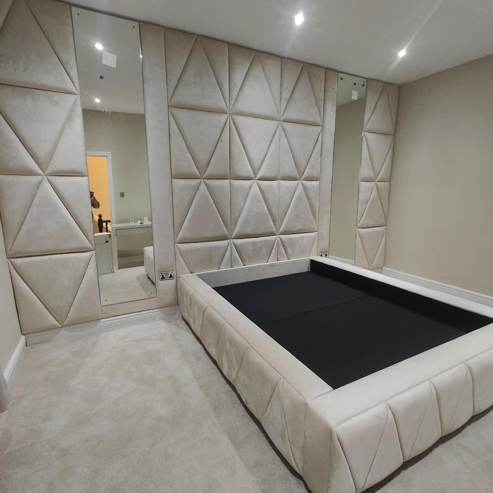 The image depicts a modern minimalist bedroom featuring a large beige upholstered bed frame with a black mattress surface. The walls are covered with plush fabric panels arranged in a geometric triangle pattern, creating a textured 3D effect. Mirrors on either side enhance the room's depth, while recessed ceiling lights provide a warm glow over the neutral-toned carpet and walls, resulting in a sophisticated ambiance.