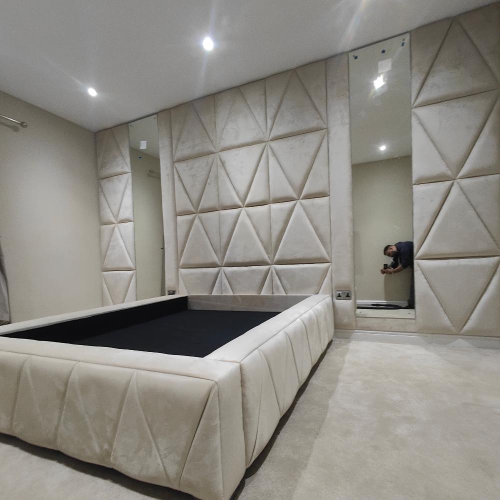 The image showcases a modern bedroom centered around a bed frame with quilted triangular patterns, matching the padded geometric wall panels. Recessed ceiling lights provide soft illumination, while a tall mirror on the right reflects part of the room and a person with a camera. The light-colored carpet complements the room’s neutral tones.