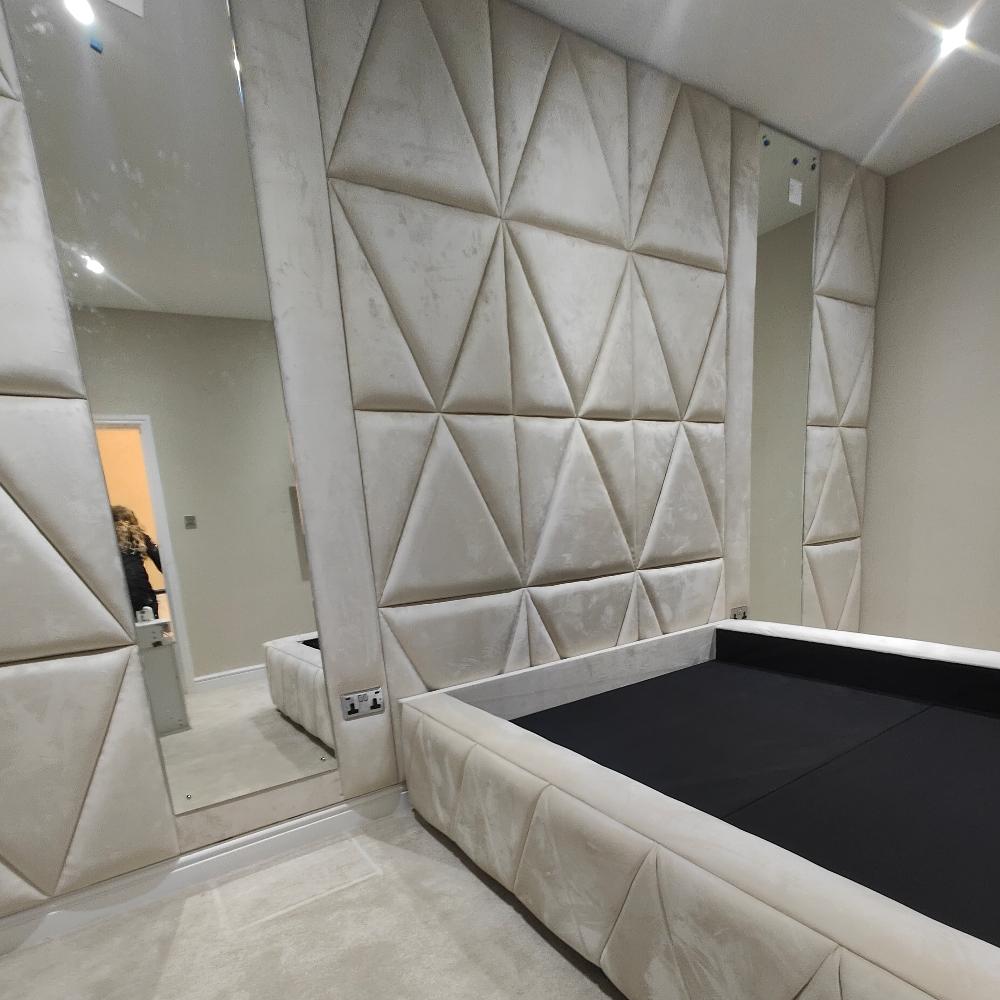 The image shows a minimalist bedroom featuring a cream upholstered bed frame and walls with quilted, triangular panels. A tall mirror on the left reflects part of the room and a silhouette at the doorway. Soft lighting and a muted color scheme create a cozy, contemporary atmosphere—perfect for The Temptress Wall Panel Bed.